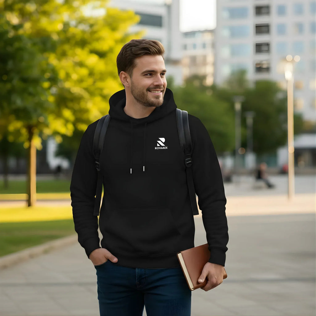 Man wearing a black Rovarix hoodie with a logo, holding a book, walking outdoors.