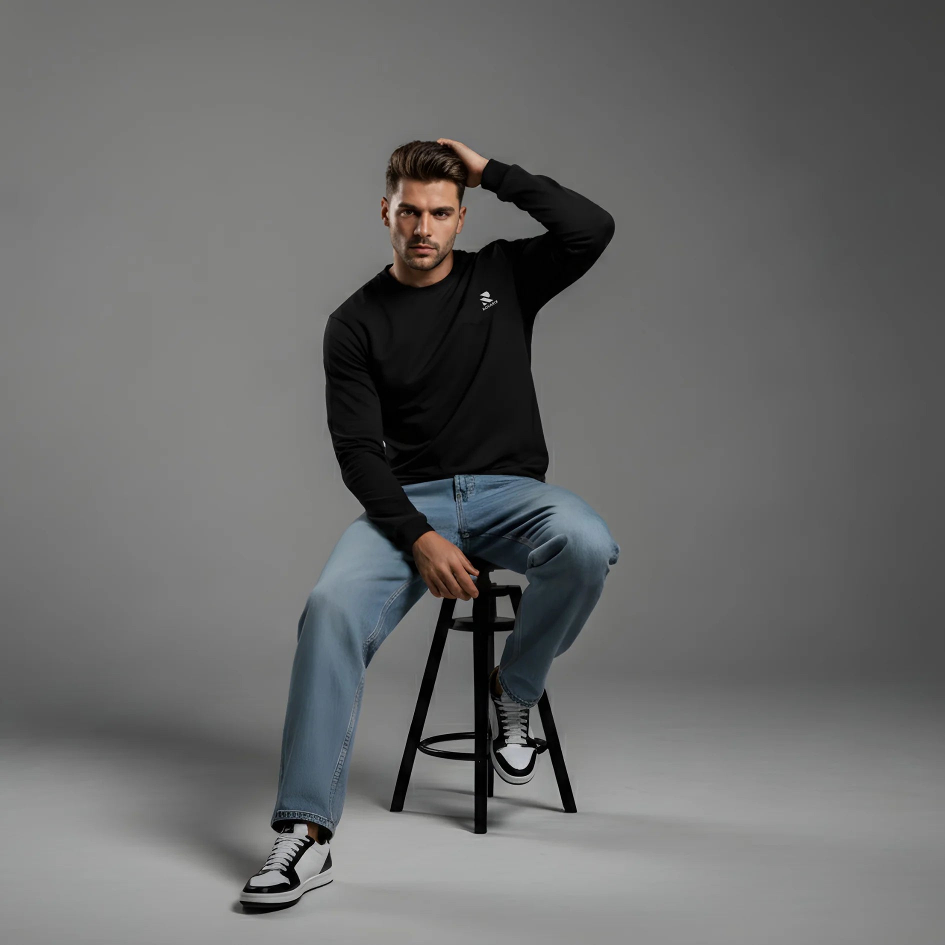 Man sitting on a stool wearing a black Rovarix sweatshirt, light blue jeans, and black and white sneakers against a gray background.