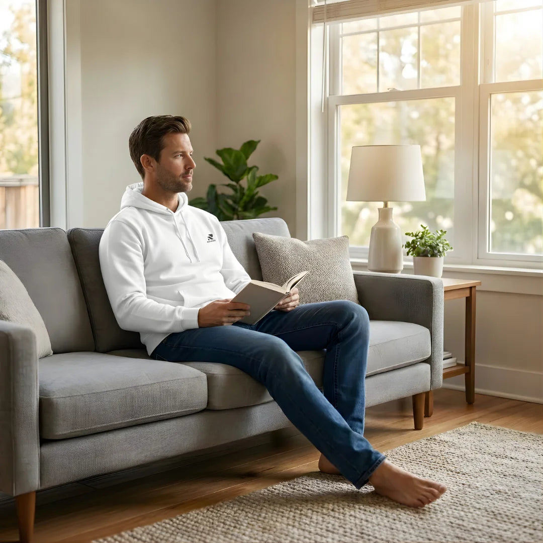 Man sitting on a couch reading a book in a bright living room.